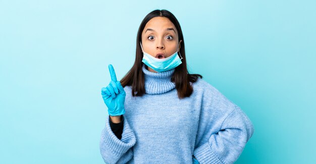 Jeune femme brune de race mixte protégeant avec un masque et des gants sur le mur bleu dans l'intention de réaliser la solution tout en levant un doigt