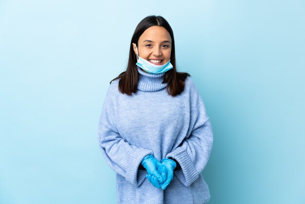 Jeune femme brune de race mixte protégeant avec un masque et des gants sur le mur bleu en riant