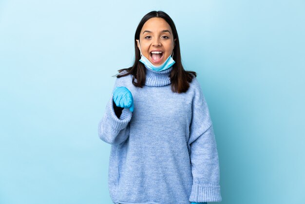 Jeune femme brune de race mixte protégeant avec un masque et des gants sur le mur bleu surpris et pointant