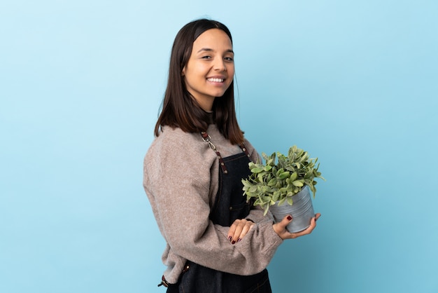 Jeune femme brune de race mixte tenant une plante sur un mur bleu isolé avec les bras croisés et impatient