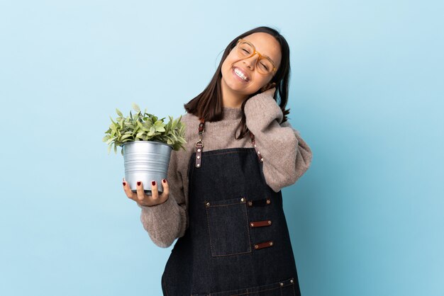 Jeune femme brune de race mixte tenant une plante sur le mur bleu en pensant à une idée.