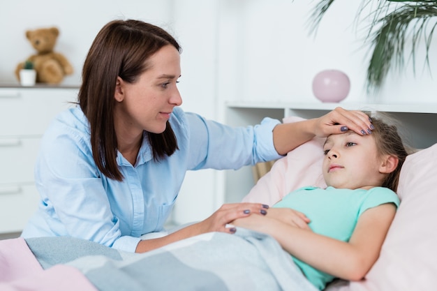 Jeune femme brune regardant sa fille malade tout en touchant le front tandis que la jeune fille couchée dans son lit