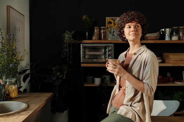 Jeune femme brune reposante en tenue décontractée tenant une tasse de thé ou de café