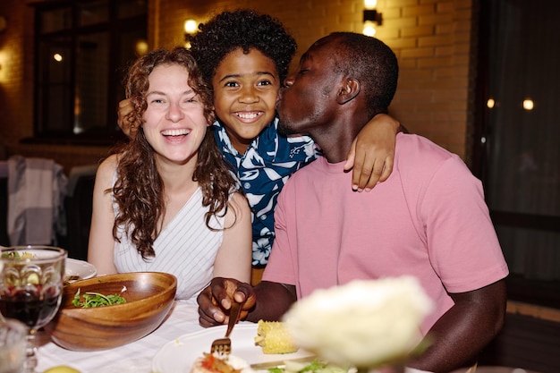 Jeune femme brune en robe blanche riant tandis qu'un garçon mignon embrassant ses heureux parents interculturels assis à table servie pendant le dîner