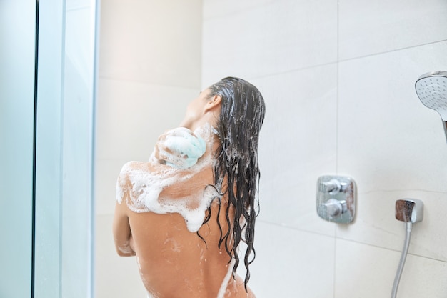 Jeune femme brune se laver le corps avec une douche en luffa