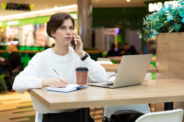 Jeune femme brune séduisante assise à une table et travaillant sur un ordinateur portable à l'aide d'un téléphone portable