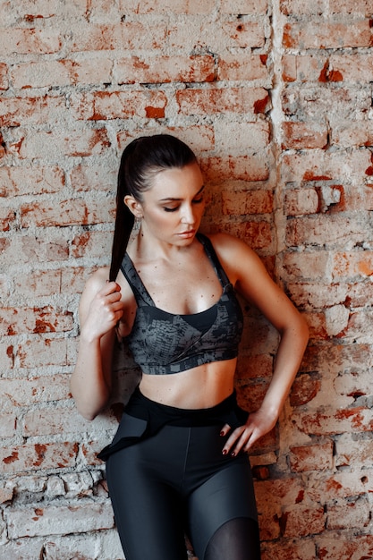 Jeune femme brune sexy en leggings de sport et haut posant contre un mur de briques dans le Studio avec style loft.