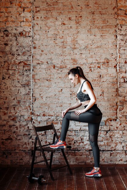 Jeune femme brune sexy en leggings de sport et haut posant contre un mur de briques dans le Studio avec style loft.