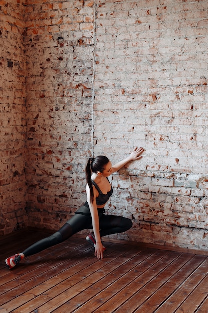 Jeune femme brune sexy en leggings de sport et haut posant contre un mur de briques dans le Studio avec style loft.