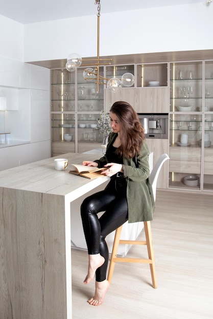 Jeune femme brune solitaire assise dans la cuisine avec une tasse de thé et un livre de lecture