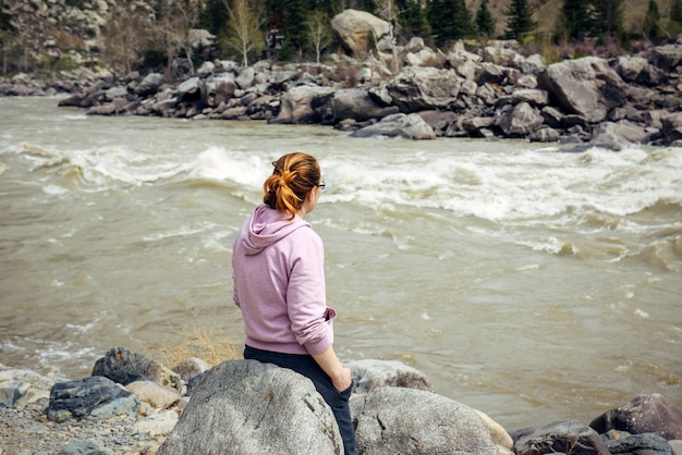 Jeune femme brune solitaire d'humeur triste assise dos à la caméra sur une grosse pierre grise près de la rivière de montagne orageuse.