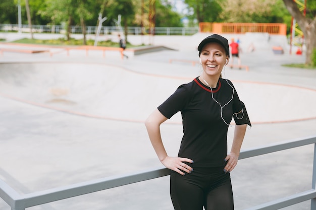 Jeune femme brune souriante athlétique en uniforme noir et casquette avec écouteurs écoutant de la musique, debout avant ou après la course, s'entraînant dans le parc de la ville à l'extérieur