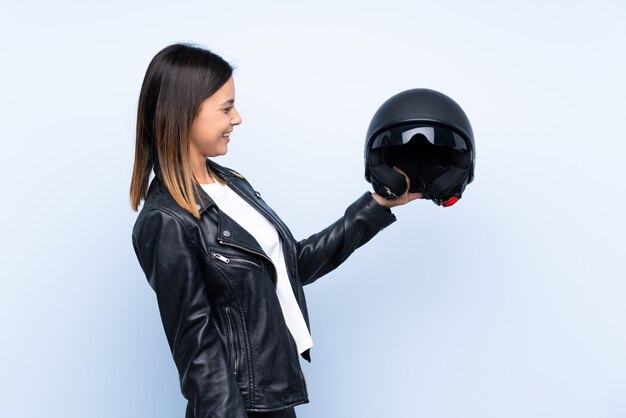 Jeune femme brune tenant un casque de moto sur un mur bleu isolé avec une expression heureuse