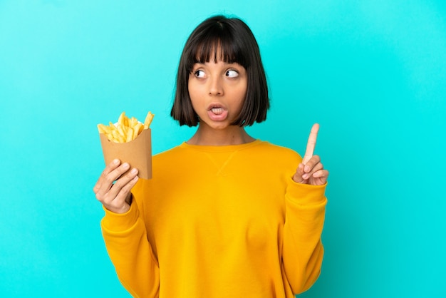 Jeune femme brune tenant des frites sur fond bleu isolé en pensant à une idée pointant le doigt vers le haut