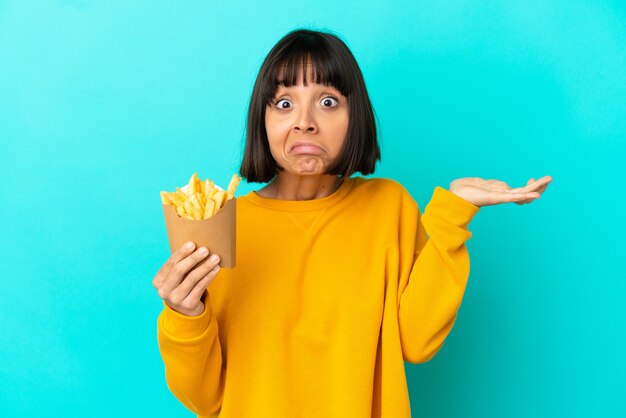 Jeune femme brune tenant des frites sur un mur bleu isolé ayant des doutes en levant les mains