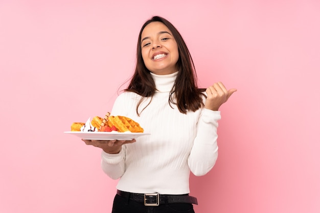 Jeune femme brune tenant des gaufres sur fond rose isolé avec le pouce en l'air et souriant