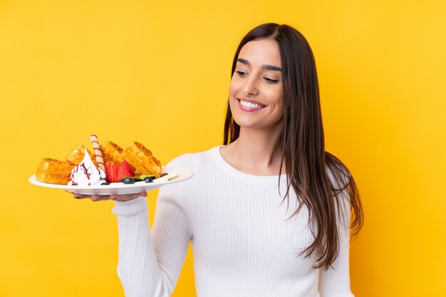 Jeune femme brune tenant des gaufres sur un mur isolé avec une expression heureuse