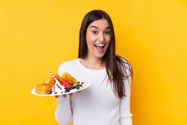 Jeune femme brune tenant des gaufres sur un mur isolé avec surprise et expression faciale choquée