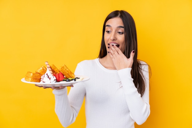 Jeune femme brune tenant des gaufres sur un mur isolé avec surprise et expression faciale choquée