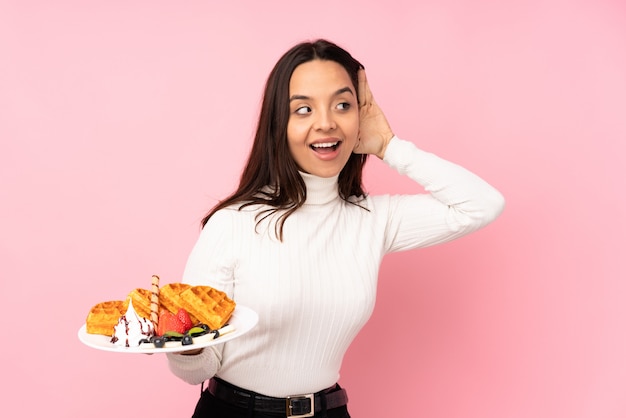 Jeune femme brune tenant des gaufres sur le mur rose en écoutant quelque chose en mettant la main sur l'oreille