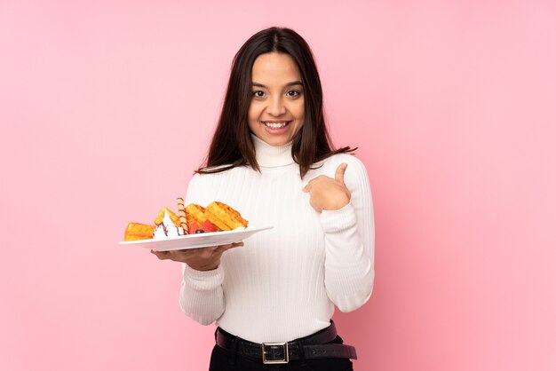 Jeune femme brune tenant des gaufres sur un mur rose isolé avec une expression faciale surprise