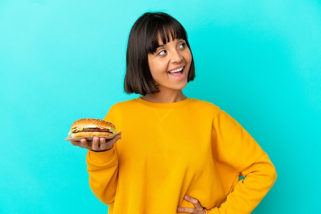 Jeune femme brune tenant un hamburger sur fond isolé en pensant à une idée tout en levant les yeux