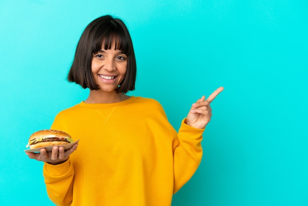 Photo jeune femme brune tenant un hamburger sur fond isolé, pointant le doigt sur le côté