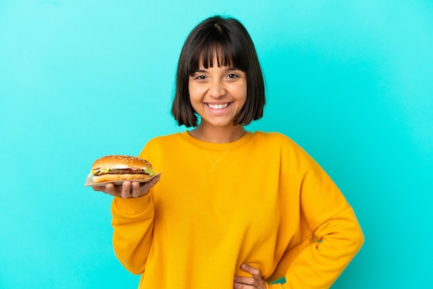 Jeune Femme Brune Tenant Un Hamburger Sur Fond Isolé Posant Avec Les Bras à La Hanche Et Souriant