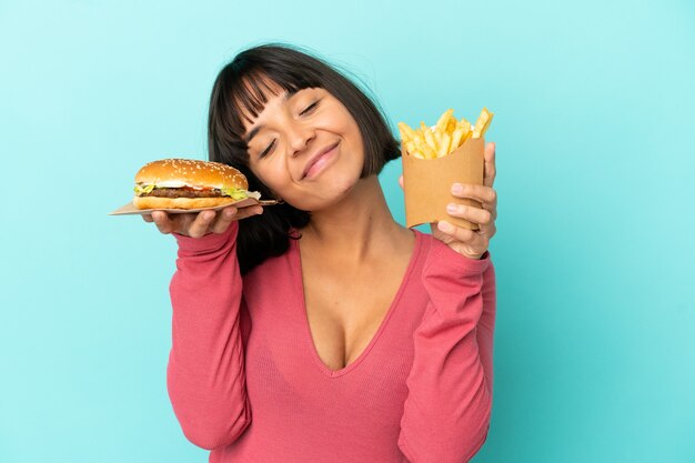 Jeune femme brune tenant un hamburger et des frites frites sur fond bleu isolé