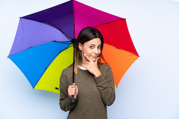 Jeune femme brune tenant un parapluie sur le mur bleu isolé en pensant à une idée