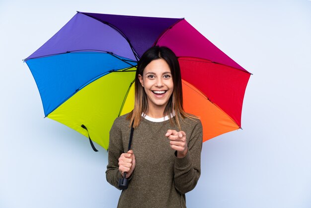 Jeune femme brune tenant un parapluie sur le mur bleu pointe le doigt vers vous