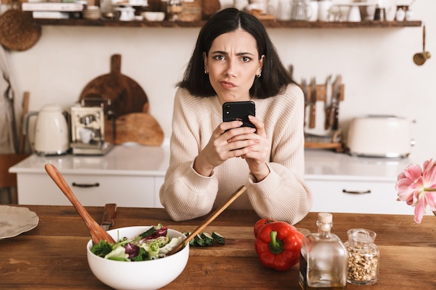 Jeune femme brune tenant le smartphone tout en cuisinant une salade verte saine avec des légumes dans une cuisine élégante à la maison