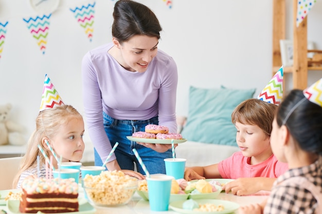 Jeune femme brune en tenue décontractée tenant la plaque avec des beignets tout en regardant un groupe de petits enfants à la maison d'anniversaire