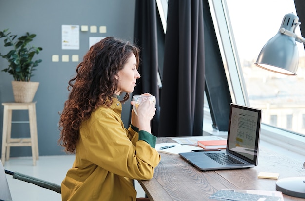 Jeune femme buvant du café assise à la table devant un ordinateur portable et travaillant à la maison