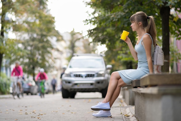 Une jeune femme buvant du café d'une tasse en papier assise sur un banc de rue de la ville dans un parc d'été