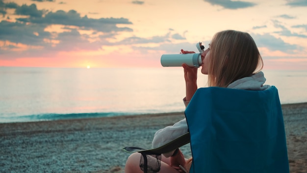 Jeune femme buvant du thermos et assise sur des chaises de camping sur la vue de profil de plage