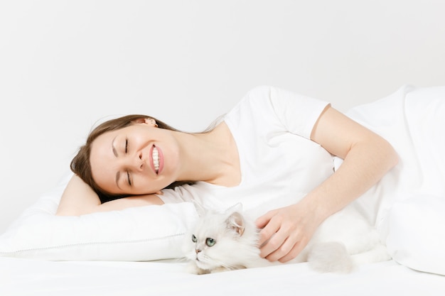 Jeune femme calme allongée dans son lit avec un mignon chat persan persan argenté, un drap, un oreiller, une couverture sur un mur blanc