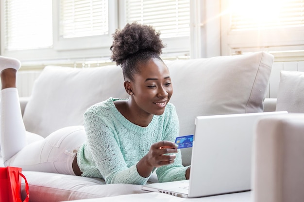 Jeune femme sur le canapé achetant en ligne avec une carte de débit Belle fille noire utilisant un ordinateur portable pour faire des achats en ligne à la maison