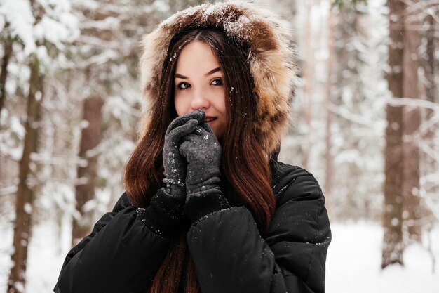 Jeune femme en capuche debout dans la forêt d'hiver se réchauffant les mains en plein air
