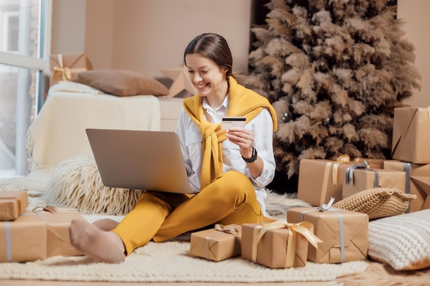 Jeune femme avec carte de crédit shopping en ligne sur ordinateur portable dans un intérieur de Noël confortable