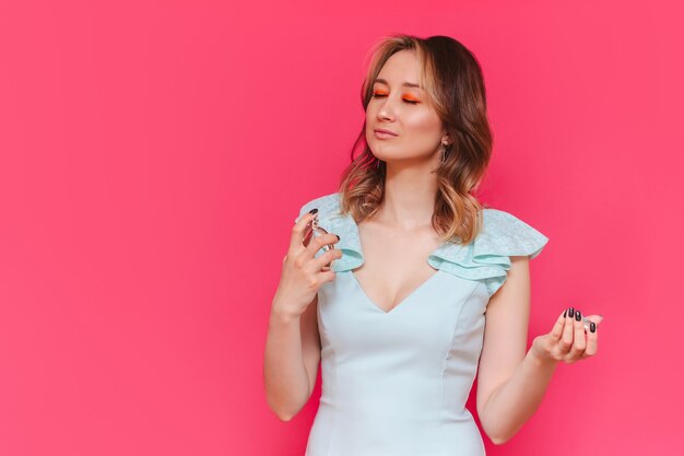 Une jeune femme caucasienne assez brune, les yeux fermés dans une robe élégante bleu clair tenant une bouteille de parfum à la main et l'appliquant sur son cou isolée sur un mur rose de couleur vive