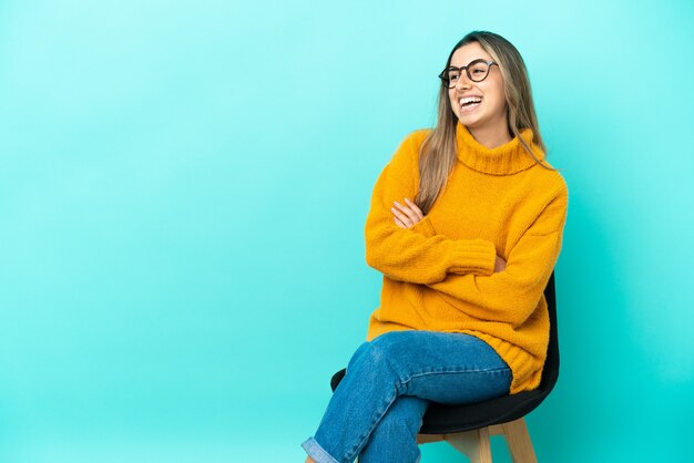 Jeune femme caucasienne assise sur une chaise isolée sur fond bleu heureux et souriant