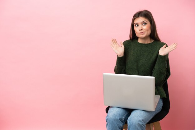 Jeune femme caucasienne assise sur une chaise avec son ordinateur portable isolé sur fond rose ayant des doutes en levant les mains