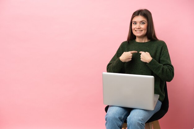 Jeune femme caucasienne assise sur une chaise avec son ordinateur portable isolé sur fond rose avec une expression faciale surprise