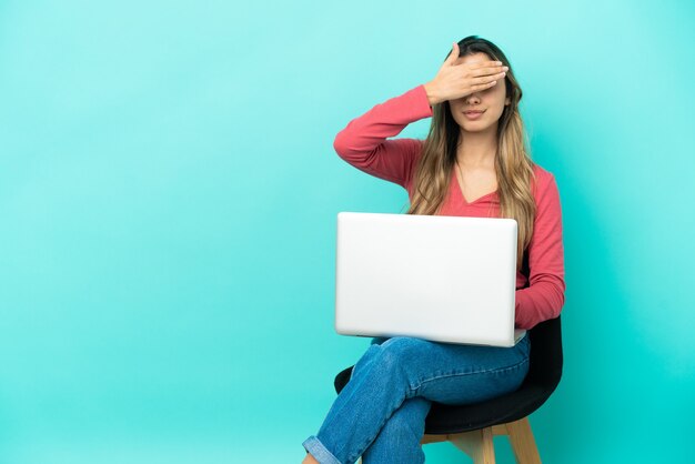 Jeune femme caucasienne assise sur une chaise avec son pc isolé sur fond bleu couvrant les yeux à la main. Je ne veux pas voir quelque chose