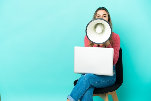 Jeune femme caucasienne assise sur une chaise avec son pc isolé sur fond bleu criant à travers un mégaphone