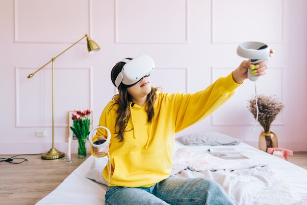 Jeune femme caucasienne assise sur le lit et utilisant un casque VR, tenant des contrôleurs et regardant en réalité virtuelle.