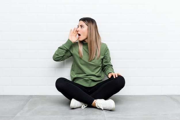 Jeune femme caucasienne assise sur le sol en criant et tenant la paume près de la bouche ouverte.