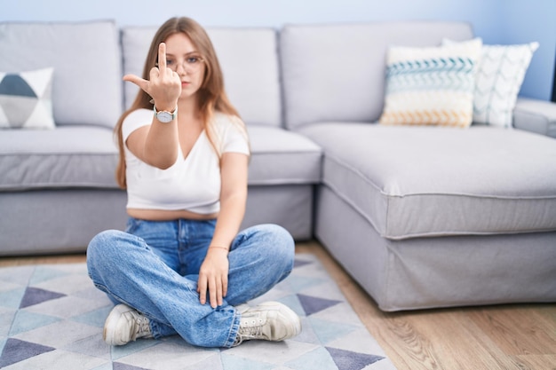 Jeune femme caucasienne assise sur le sol dans le salon montrant le doigt du milieu, expression de baise impolie et grossière