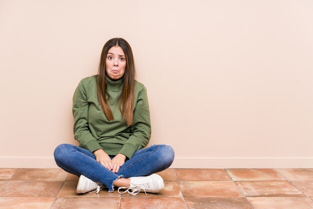 Jeune femme caucasienne assise sur le sol hausse les épaules et les yeux ouverts confus.